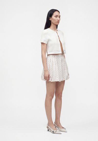 Femme debout portant un cardigan court blanc boutonné, une mini-jupe plissée à motifs, et des chaussures blanches pointues à talons bas.