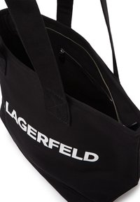 Sac fourre-tout en toile noire avec texte "LAGERFELD" blanc contrastant, doté de bretelles réglables et d'une poche intérieure zippée.