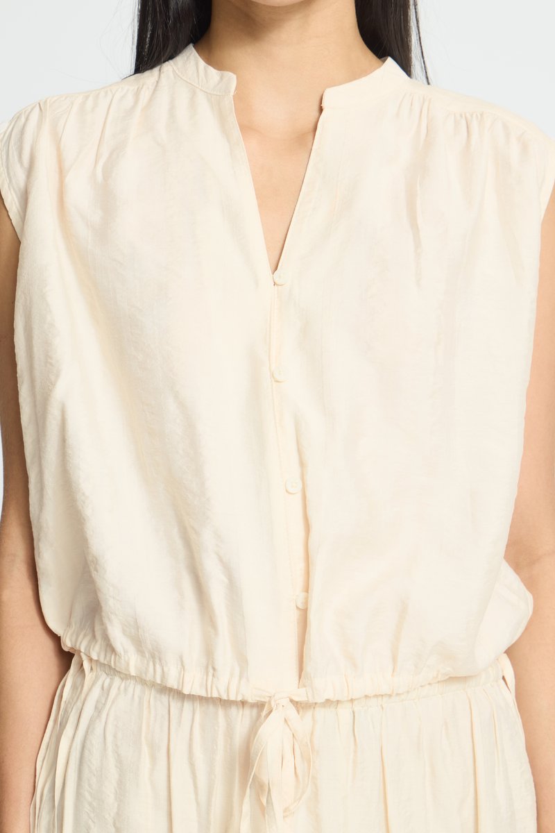 Femme portant un haut crème sans manches boutonné avec une taille à cordon et des détails de tissu froncé.