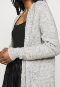 Ljusgrå, mjuk stickad cardigan med öppen framsida och långa ärmar, med ribbad finish på manschetterna. Bärs över en svart topp.