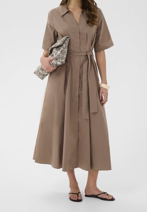 Donna che indossa un vestito midi marrone con maniche corte, tiene in mano una pochette in pelle di serpente e indossa sandali neri bassi e un braccialetto bianco.