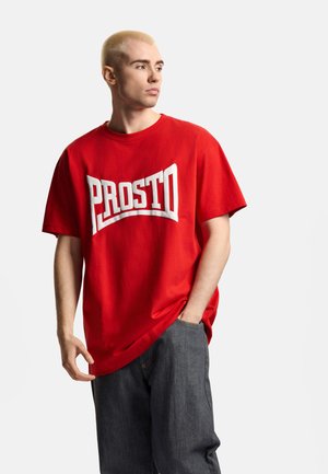 Blond mężczyzna w luźnej czerwonej koszulce z napisem "PROSTO" i ciemnoszarych spodniach, patrzący w bok z jedną ręką w kieszeni.