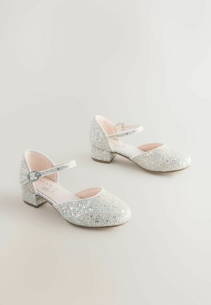 Spīdīgas sudraba kurpes ar apaļu purngalu, potītes siksniņu un zemu bloku papēdi. Augšdaļa ir ar spīdīgu tekstūru un gludu rozā oderi.