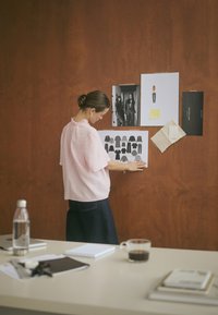 Light pink textured blouse with ruffled neckline and dark denim pants. White and black designs pinned on a dark brown wall. Desktop items visible.