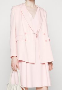 Dvouřadový blazer v světle růžové barvě s klopami, velkými knoflíky a bočními kapsami, nošený přes shodné růžové šaty. Držená texturovaná kabelka.