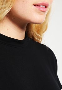 Černý top s vysokým límcem, hladkou texturou, přiléhavým střihem a minimalistickým stylem, který se vyznačuje čistými liniemi a bez viditelného kovového prvku.