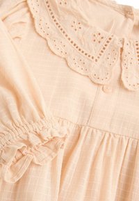 Robe couleur pêche avec un motif en carrés texturé. Dispose d'un col en dentelle en forme de scallop, de manches bouffantes avec élastique et d'une fermeture à boutons.