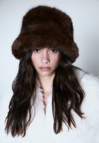 Donna con lunghi capelli scuri che indossa un grande cappello di pelliccia marrone scuro e un cappotto di pelliccia bianca, che guarda direttamente in camera con un'espressione neutra.