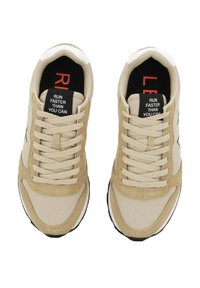 Beige Sneaker aus Stoff und Wildlederobermaterial, ausgestattet mit Schnürsenkeln, einem auffälligen schwarzen Etikett mit rotem Text und einer schwarzen Außensohle für Traktion.