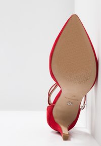 Chaussure à talon kitten en daim rouge, à bout pointu, tenue sur son talon, montrant la semelle texturée et la bride de cheville avec boucle.