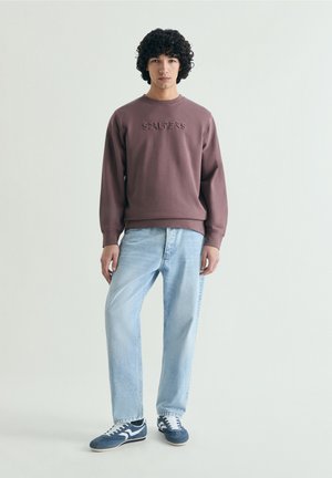 Jonge man met krullend haar die een mauve sweatshirt, lichtblauwe spijkerbroek en blauw-witte sneakers draagt, staand tegen een effen lichte achtergrond.