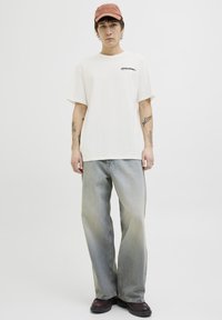 Beige t-shirt met korte mouwen en zwart logo, gecombineerd met lichtblauwe losse jeans en bruine schoenen. Aangevuld met een roze pet.