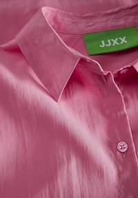 Camicia rosa con colletto in tessuto liscio; presenta un'etichetta verde con "JJXX" e una chiusura con un solo bottone. Leggera lucentezza e trama fine.