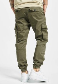 Olivové nohavice s elastickými manžetami, viacerými bočnými vreckami a zapínacími zadnými vreckami, skombinované s bielymi teniskami.