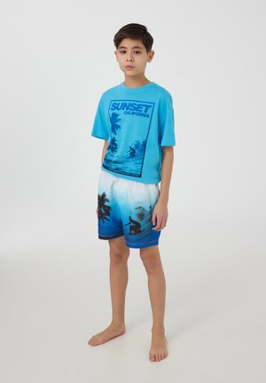 Maglietta grafica blu con stampa "SUNSET CALIFORNIA" e design per il surf, abbinata a pantaloni da bagno blu a fantasia con palme e surfisti.