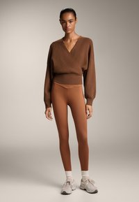 Pulover maro din tricot texturat, asortat cu colanți maro ajustați. Modelul poartă adidași deschiși la culoare cu șosete albe. Fundal neutru.