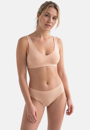 Donna con capelli biondi che indossa un completo coordinato di reggiseno beige senza cuciture e mutandine a vita alta su uno sfondo chiaro e uniforme.