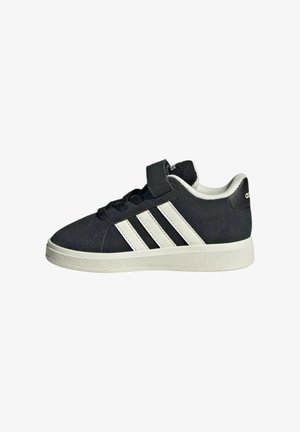 Zapatillas de ante negro con rayas blancas, una correa de velcro y suela de goma. El diseño tiene un perfil bajo y detalles texturizados.