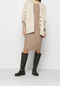 Cardigan en tricot beige avec de grands boutons sur une robe beige à côtes. Bottes en cuir noires jusqu'au genou avec une finition texturée et des semelles épaisses.
