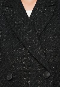 Crni dvoredni kaput napravljen od teksturiranog materijala s suptilnim sjajem, s ovratnikom i dva crna dugmeta kao hardverskim akcentima.