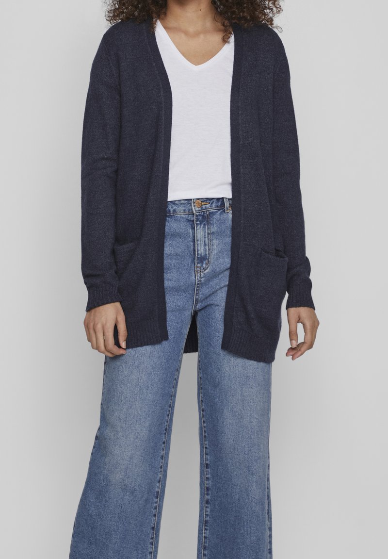 Femme portant un cardigan bleu marine ouvert sur le devant, un t-shirt blanc à col en V et un jean taille haute bleu clair à jambes larges.