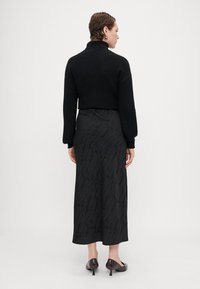 Melnas adītas īsās lijas džemperis ar augstu apkakli, kombinēts ar garu melnu kleitu ar strukturētu rakstu. Uzvilktas tumšas, spīdīgas kurpes ar augstu papēdi.