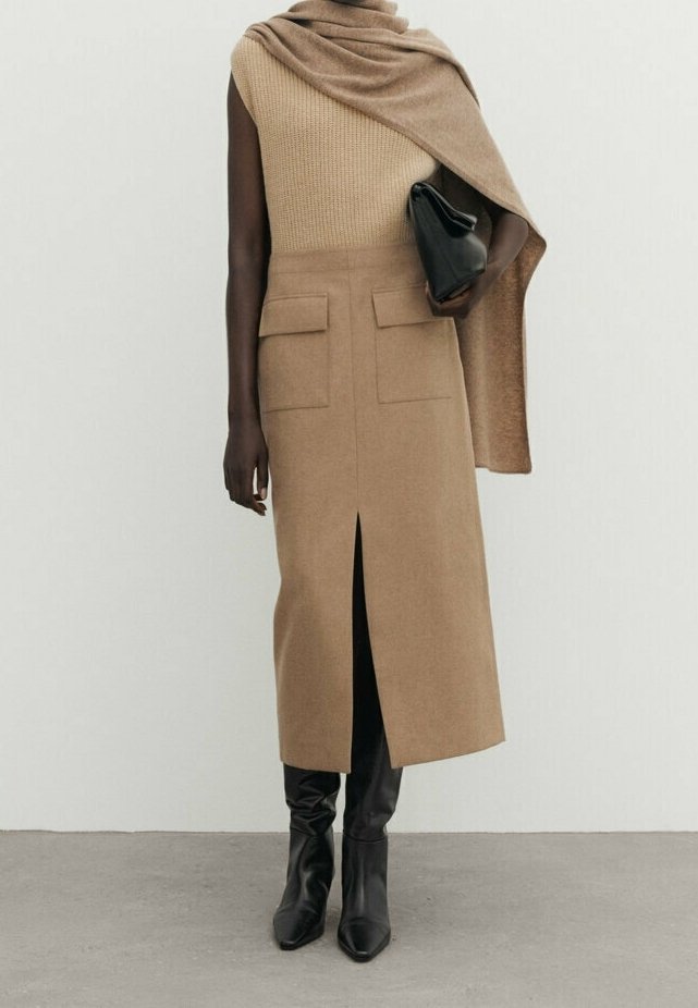 Femme portant un haut en tricot beige sans manches avec une jupe assortie comportant une fente devant et des poches, bottes noires, tenant une pochette noire, châle beige sur l'épaule.