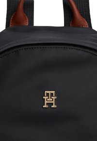 Black fabric backpack with brown leather accents and a gold metal logo featuring stylized interlocking letters on the front.