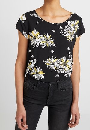 Personne portant une chemise noire à manches courtes avec un motif floral jaune et blanc, associée à un jean noir taille haute, debout.