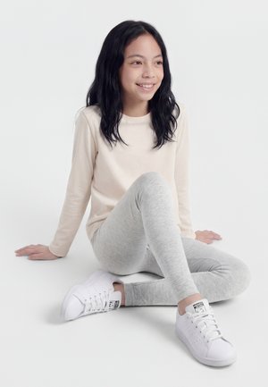 Ung jente med langt svart hår som sitter med beina i kors, iført en kremhvit langermet skjorte, grå leggings og hvite joggesko, smilende.