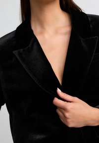 Schwarzer Samtblazer mit Reverskragen und tiefem V-Ausschnitt. Der Stoff hat eine weiche, glatte Textur. Detail einer Hand, die den Revers hält.