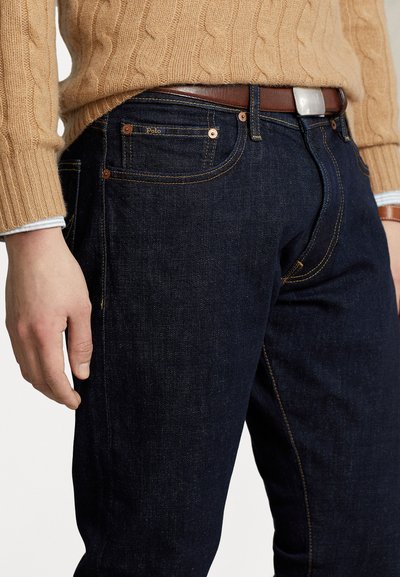 Jeans din denim de culoare albastru închis au o croială slim, cusături contrastante și o curea din piele cu o cataramă argintie. Țesătura are o textură netedă.