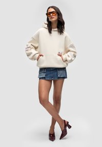 Kremowa bluza z kapturem o oversize'owych rękawach, z kieszeniami z przodu i haftowanym detalem. połączona z dżinsową mini spódnicą i burgundowymi butami na obcasie.