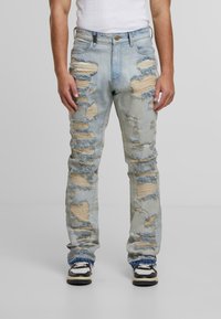 Šviesiai mėlynos nusidėvėjusios džinsinės kelnės su dideliais įplyšimais, ištrintais kraštais ir penkių kišenių dizainu, derinamos su juodai baltais sportiniais batais.