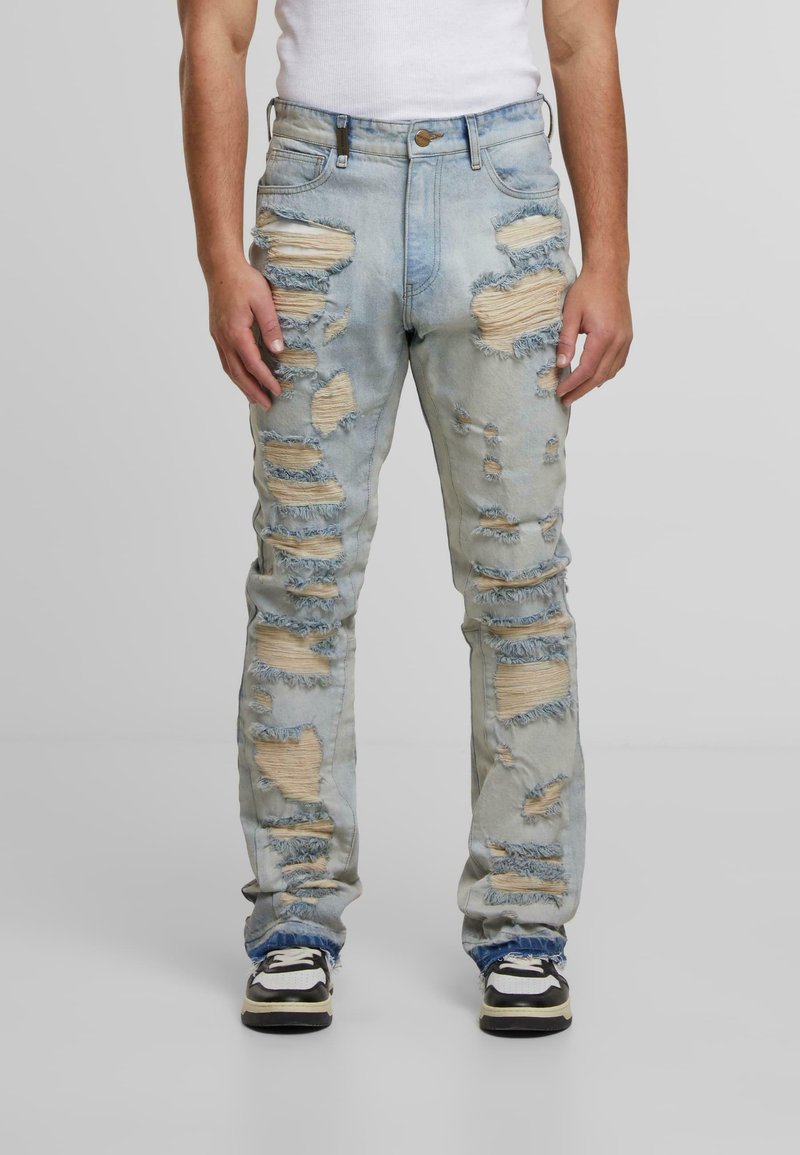 Šviesiai mėlynos nusidėvėjusios džinsinės kelnės su dideliais įplyšimais, ištrintais kraštais ir penkių kišenių dizainu, derinamos su juodai baltais sportiniais batais.