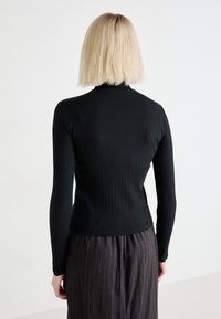 Svart ribbad långärmad topp med hög krage, figursydd design och slät textur. Bärs med grå randiga byxor, sedd bakifrån.