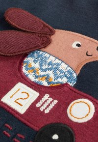 Hímzett anyagon egy barna nyúl látható, nagy fülekkel, kék és narancssárga mintás pulóverben, valamint egy bordó autó fehér díszítésekkel.