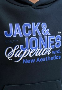 Sudadera con capucha negra con texto en azul y blanco audaz: "JACK & JONES", "Superior wrd" y "New Aesthetics". El material parece suave y de corte regular.