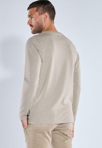 Beige langärmliges Shirt mit Rundhalsausschnitt und subtilen Nahtdetails, kombiniert mit hellen Hosen. Einfache, weiche Stofftextur.