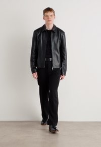 Veste en cuir noir, fermeture éclair, col montant; portée sur une chemise noire et un pantalon; chaussures noires avec un léger talon; fond neutre.