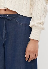 Midsection of person wearing a cream cable-knit sweater and blue drawstring pants, hand relaxed by side with tattoos on fingers and wrist.