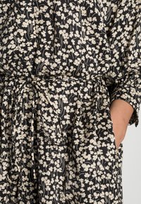 Schwarzes Stoffkleid mit weißem Blumenmuster, tailliert mit einem Band. Verfügt über lange Ärmel und seitliche Taschen. Strukturierte Optik.