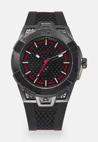 Reloj con correa de goma negra, esfera negra texturizada, acentos rojos y herrajes plateados. Presenta una caja audaz y marcadores de hora elevados.