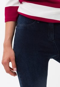 Jeans de mezclilla en azul oscuro con un diseño ajustado y una textura suave, combinados con una blusa a rayas rojas y blancas. Mano descansando en la cadera.