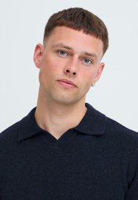 Fiatal férfi, rövid barna hajjal és kék szemekkel, sötét tengerészkék pulóvert visel, kissé megdöntött fejjel, semleges arckifejezéssel, világos háttér előtt.