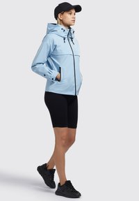Veste imperméable bleu clair avec une capuche, fermeture éclair devant et accents noirs. Associée à des shorts noirs côtelés et des chaussures de sport noires.