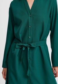 Grünes langärmliges Kleid mit V-Ausschnitt, durchgehender Knopfleiste und Taillengürtel. Hergestellt aus glattem Stoff in einheitlicher Farbe ohne Muster.