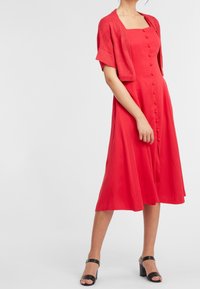Robe midi rouge avec encolure carrée, boutons sur le devant et cardigan en tricot court. Présentant une texture lisse et portée avec des sandales à talons noires.