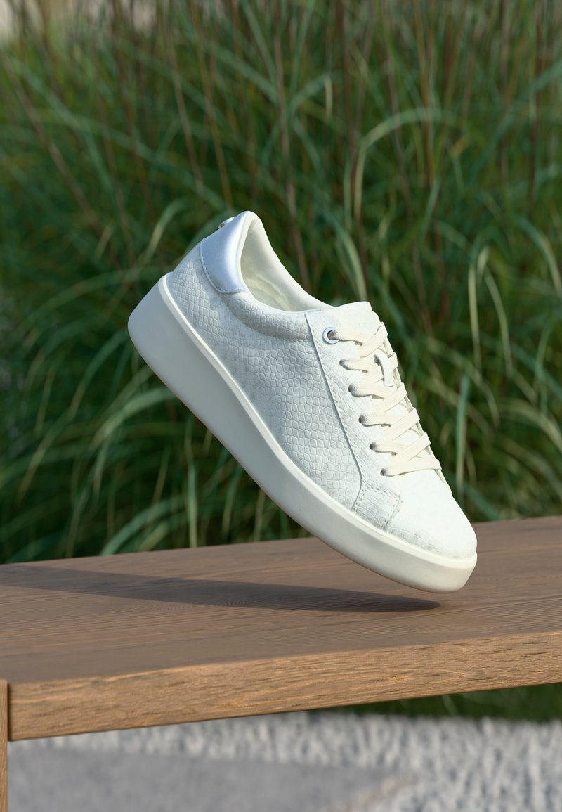 Baskets blanches texturées avec lacets flottant au-dessus d'un banc en bois, hautes herbes vertes floues en arrière-plan en plein air.