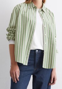 Camisa de botones a rayas verdes y blancas con un bolsillo, hecha de una tela ligera; usada sobre una camiseta blanca y combinada con jeans oscuros.